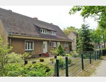 Ein einstöckiges Wohnhaus mit brauner Putzfassade und Satteldach, umgeben von einem kleinen Garten mit Pflanzkübeln und Sträuchern. Im Vordergrund steht ein Metallzaun, und rechts führt ein gepflasterter Gehweg entlang.