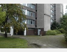 Mehrstöckiges Wohngebäude aus Beton mit großen Fenstern, flankiert von Bäumen und Sträuchern. Im Vordergrund ein gepflasterter Weg neben einer Rasenfläche und einem Baum mit orangefarbenen Beeren.