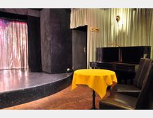 Bühnenbereich mit schwarzem Klavier und einer kleinen Sitzgelegenheit mit Tisch und gelber Tischdecke im Vordergrund, daneben ein verhängter Vorhang aus metallisch glänzendem Stoff. Im Hintergrund sind grüne Vorhänge, eine Tür und ein Hinweis auf eine Toilette sichtbar.