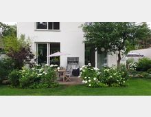 Moderne Hausfassade mit weißem Putz und großen Glas-Schiebetüren, die zu einer Terrasse führen. Die Terrasse ist mit einem Gartentisch, Stühlen, einem Strandkorb und einem rosa Sonnenschirm möbliert, umgeben von Blumenbeeten mit weißen Hortensien und einem Baum mit hängenden Lampions im gepflegten Garten.