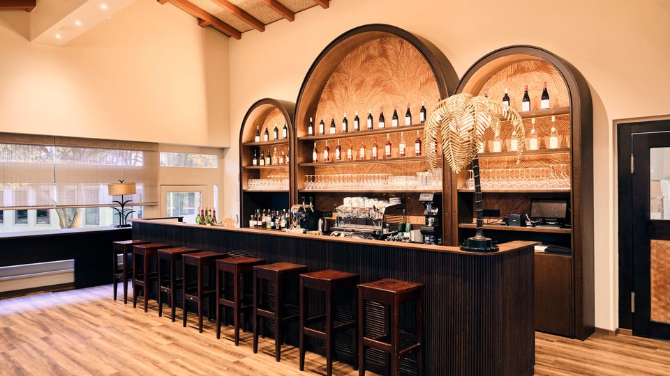 A modern bar interior with a wooden counter lined with high stools and an arched shelving unit displaying bottles and glassware. The warm space features a patterned backdrop, wooden ceiling beams, and a decorative golden fern centerpiece.
