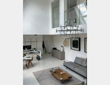 Modernes, helles Wohnzimmer mit hohen Decken, weiß gestrichenen Wänden und minimalistischer Einrichtung. Der Raum zeigt eine graue Couch, einen rechteckigen Holztisch, eine große Designerlampe und eine offene Galerie mit Glasgeländer im oberen Bereich.