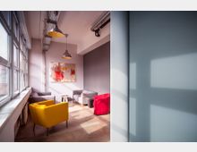 Heller, moderner Loungebereich mit einer Fensterfront auf der linken Seite, mehreren farbigen Sesseln (gelb, rot und grau), einem kleinen Beistelltisch und abstrakter Wandkunst. Zwei Hängeleuchten und sichtbare Kabelträger an der Decke ergänzen die industrielle Gestaltung.