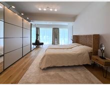 Helles Schlafzimmer mit einem Bett mit geflochtenem Kopfteil, Holz-Nachttisch und großer Spiegelschrankwand links. Der Raum hat einen Holzfußboden, einen Teppich und eine Fensterfront mit halbtransparenten Vorhängen, die Tageslicht hereinlassen.