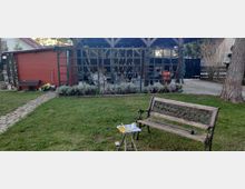 Ein Garten mit einer Holzbank und einem kleinen Tisch im Vordergrund. Im Hintergrund befinden sich ein rotes Gartenhaus, ein größerer Unterstand mit Holzrahmenkonstruktion und Gartenutensilien sowie ein gepflasterter Weg.