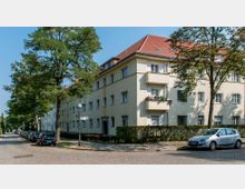 Wohnhaus in einer ruhigen Wohngegend mit heller Fassade und roten Ziegeldächern, flankiert von Bäumen. Links führt eine gepflasterte Straße mit parkenden Autos entlang, umgeben von gepflegten Hecken.