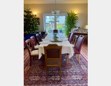 Festlich gedeckter Esstisch in einem Wohnraum mit weißem Tischtuch, Geschirr und Gläsern, umgeben von Holzstühlen. Im Hintergrund stehen ein geschmückter Weihnachtsbaum und eine große Zimmerpflanze neben einer Glastür mit Blick auf einen Garten.