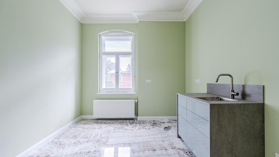 Heller, leerer Küchenraum mit hellgrünen Wänden, hoher Decke mit Stuckleisten und einem großen Fenster über einem Heizkörper. Rechts steht eine moderne Küchenzeile mit grauer Arbeitsplatte, Spülbecken und Wasserhahn; der Boden ist mit glänzenden marmorierten Fliesen ausgelegt.