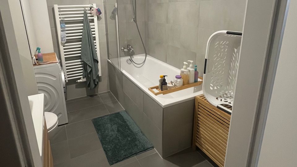 Modernes Badezimmer mit grauen Boden- und Wandfliesen, Badewanne mit Glas-Duschabtrennung und Handbrause sowie Handtuchheizkörper an der Stirnwand. Links stehen Waschmaschine und Waschbecken, rechts ein Holzschrank mit Wäschekorb; ein kleiner Teppich liegt vor der Wanne.