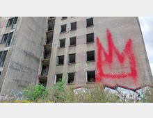 Fassade eines verlassenen, mehrstöckigen Gebäudes mit leeren Fensterrahmen und sichtbaren Verwitterungsspuren. An der Wand ist ein großes rotes Graffiti in Form einer Krone zu erkennen, umgeben von überwuchertem Gestrüpp im Vordergrund.