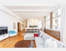 Heller, offener Loft-Wohnraum mit Küche und Essbereich, weißen Deckenbalken und Holzboden. Große Fensterfront mit freigelegter Ziegelwand sowie moderne Einrichtung mit Sofa, Couchtisch und Kücheninsel.