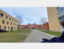 Ein gepflasterter Weg führt durch eine grüne Wiese zu einem großen Backsteingebäude mit symmetrischen Fassaden und zahlreichen Fenstern. Auf beiden Seiten des Bildes stehen modernere Gebäude aus hellen Ziegelsteinen, und im Hintergrund sind kahle Bäume sichtbar.