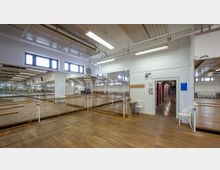 Ein heller Tanz- oder Übungsraum mit Holzboden, großen Wandspiegeln und Ballettstangen. An der rechten Seite befindet sich ein Durchgang zu einem Flur, und die Deckenbeleuchtung sowie Fenster sorgen für gleichmäßiges Licht.