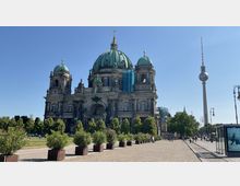 Berliner Dom mit großer grüner Kuppel, flankiert von zwei kleineren Türmen, im Vordergrund ein gepflasterter Platz mit Pflanzen in Kübeln. Im Hintergrund ist der Fernsehturm zu sehen, rechts befinden sich Straßenlaternen und eine Straßenbahnhaltestelle.