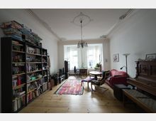 Helles Wohnzimmer mit Holzdielenboden, einem großen Bücherregal an der linken Wand und einer roten Couch auf der rechten Seite. In der Mitte befindet sich ein bunter Teppich, ein runder Tisch und Sessel; am Fenster steht ein Schreibtisch mit Bürostuhl.
