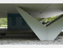 Eine Detailansicht eines modernen Gebäudes mit großen, V-förmigen Stützpfeilern aus Beton. Im Hintergrund ist eine mit blauen Fliesen verkleidete Wand sowie Grünflächen mit Bäumen sichtbar.