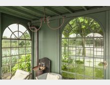 Innenraum eines kleinen grünen Pavillons mit großen, bogenförmigen Fenstern, die den Blick auf eine Gartenlandschaft mit Bäumen und Sträuchern ermöglichen. Im Raum stehen ein rustikaler Ofen, ein Metalltisch mit Wasserhahn sowie ein Korbsessel mit Kissen.