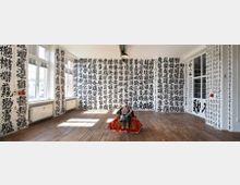 Ein heller Raum mit weißen Wänden, die vollständig mit großformatigen schwarzen chinesischen Schriftzeichen bedeckt sind. Auf dem Holzboden steht zentral eine bunt dekorierte Figur auf einem roten Tuch; an den Wänden befinden sich große Fenster und Heizkörper.