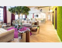 Ein modernes Büro mit einer gemütlichen Sitzecke, bestehend aus beigen Sofas, gemusterten Sesseln und einem Glastisch auf einem lilafarbenen Teppich. Eine grüne Mooswand, eine Glaswand mit Tür sowie Pflanzen und beleuchtete Regale prägen den Raum.