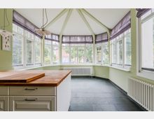 Heller Wintergarten-ähnlicher Raum mit vielen bodentiefen Fenstern rundum, hellgrünen Wänden und sichtbaren Deckenstreben unter einem spitz zulaufenden Dach. Vorne steht eine Kücheninsel mit Holzarbeitsplatte; entlang der Wände sind Heizkörper und graue Fliesen am Boden.