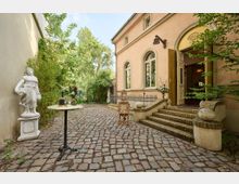 Seitlicher Eingang eines historischen Gebäudes mit aprikosenfarbener Fassade, geschwungenen Treppen aus Ziegelsteinen und hohen Fenstern. Davor eine gepflasterte Fläche mit einer weißen Statue, einem kleinen Tisch mit Champagner sowie umgebender grüner Gartenvegetation.