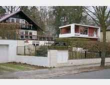 Straßenszene mit zwei Wohnhäusern: Links ein zweistöckiges Haus mit dunkler Holzfassade, Balkon und Satteldach, rechts ein modernes, zweistöckiges Gebäude mit Flachdach und roter Fassade. Vordergrund zeigt eine Steinmauer mit Metallzaun und Gehweg.