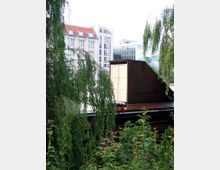 Ein mit Pflanzen und hängenden Zweigen umgebenes altes Transportfahrzeug mit einer gelben Tür, im Hintergrund moderne und historische Gebäude mit roten und gläsernen Fassaden.