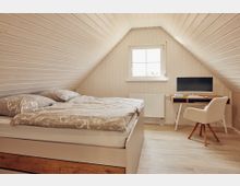 Schlafzimmer unter einer Dachschräge mit hellen Holzvertäfelungen an Wänden und Decke. Links steht ein Bett mit gemusterter Bettwäsche, rechts ein Schreibtisch mit Monitor und einem gepolsterten Stuhl, vor einem Fenster mit Tageslicht.