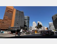 Straßenszene am Potsdamer Platz in Berlin mit modernen Hochhäusern, darunter ein orangefarbener und ein gläserner Turm. Im Vordergrund befinden sich ein kleiner Markt, Werbeplakate und der Schriftzug „Bahnhof Potsdamer Platz“.