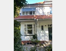 Zweistöckiges Wohnhaus mit weißer Fassade, rotem Ziegeldach und blau-weiß gestreifter Markise über einem oberen Fenster. Im Vordergrund sind ein gepflegter Garten mit Sträuchern sowie ein gepflasterter Eingangsbereich mit Treppe sichtbar.