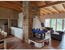 Wohnraum mit offenen Holzbalkendecken, einer gemauerten Wand aus Natursteinen und einem Kamin mit blauen Fliesen. Im Hintergrund befindet sich eine Sitzecke mit Ledersofas vor großen Fenstern, die einen Blick auf einen Garten ermöglichen.