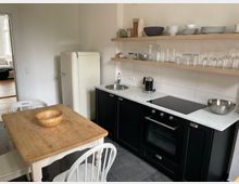 Kleine, modern eingerichtete Küche mit weißen Wänden und dunklen Unterschränken. Ein Holztisch mit Stühlen steht neben einem cremefarbenen Kühlschrank, während offene Holzregale Geschirr und Gläser über einer Arbeitsplatte mit Spüle, Kochfeld und Backofen präsentieren.