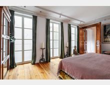 Schlafzimmer mit Holzboden, bodentiefen Fenstern mit weißen Jalousien und grünen Vorhängen. Ein großes Holzbett, dekorative Kerzenständer und eine sitzende Buddha-Statue prägen den Raum, daneben eine geöffnete zweiflügelige Holztür.