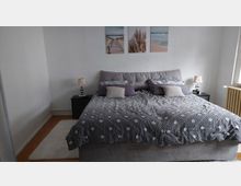 Schlichtes Schlafzimmer mit großem Doppelbett in grauer Bettwäsche, zwei Nachttischen mit Lampen und drei Wandbildern über dem Kopfteil. Rechts ein Fenster mit Heizkörper darunter, Holzboden und heller Teppich vor dem Bett.