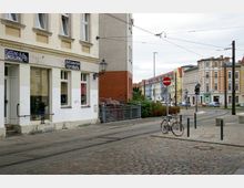 Städtische Straßenszene mit Kopfsteinpflaster, Straßenbahngleisen und Gebäuden im Hintergrund, darunter ein Geschäft mit den Schildern "Tattoo & Piercing" und "Plottarbeiten & Werbung". Im Vordergrund steht ein Fahrrad an einem Verkehrsschild angeschlossen.