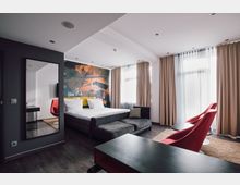 Modern eingerichtetes Hotelzimmer mit großem Doppelbett, Wand mit farbigem Graffiti-Motiv, bodenlangen Vorhängen und hellen Fenstern. Im Vordergrund zwei rote Sessel, ein dunkler Tisch und ein Teppich auf holzartigem Bodenbelag.