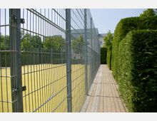 Ein umzäunter Sportplatz mit gelbem Bodenbelag, flankiert von hohen Metallgittern und einer Hecke. Im Hintergrund sind Bäume sowie ein Gebäude mit klassischem Architekturstil sichtbar.