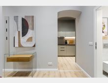 Moderner Wohnungsflur mit hellem Holzboden, einer Glas-Konsole mit Holzschubladen und abstraktem Wandbild. Ein Rundbogen-Durchgang führt in eine kompakte, graue Einbauküche; rechts ist ein offener Türrahmen zu einem Badezimmer erkennbar.