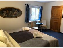 Gemütliches Hotelzimmer mit Doppelbett, auf dem gefaltete Handtücher liegen, ein kleines Fenster mit blauen Vorhängen, ein runder Tisch mit zwei Stühlen sowie eine Holztür. An der Wand hängt ein ovales Gemälde mit goldfarbenem Rahmen.