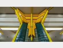 Nahaufnahme einer Säule in einer U-Bahn-Station mit grünen Fliesen und auffälligen gelben, geometrischen Gestaltungselementen, die sich bis zur Decke erstrecken. Die Decke zeigt helle Beleuchtung und strukturierte Streben im Hintergrund.
