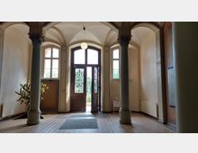 Eingangsbereich eines historischen Gebäudes mit hohen Decken, verzierten Säulen und einem großen Holztürportal mit schmiedeeisernen Ornamenten und Glaseinsätzen; dahinter ist ein Garten sichtbar. Der Boden ist mit Fliesen gestaltet, und an den Wänden befinden sich Dekorationselemente und eine Infotafel.