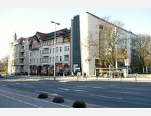 Historische Wohngebäude im Stil der Gründerzeit mit roten Dächern und Zierelementen neben einem modernen Gebäude mit Glasfassade und einem gebogenen, weißen Seitenteil, auf dem "Café Scala" steht. Davor eine breite Straße mit Fußgängerzone, Bushaltestelle und vereinzelten Passanten.