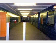 Ein unterirdischer Fußgängertunnel mit blau gefliesten Wänden, einer weißen Decke und Reihen von Deckenleuchten. Rechts befinden sich geschlossene Schalterfenster, links ein orangefarbener Kioskautomat, und im Hintergrund führen Treppen nach oben.