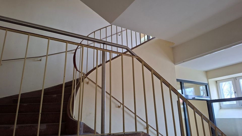 Innenansicht eines Treppenhauses mit gewendelten Treppenläufen und hellen Wänden, eingefasst von einem Metallgeländer. Große Fensterflächen lassen Tageslicht einfallen und geben den Blick nach außen frei.