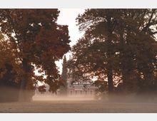 Historisches Gebäude mit Säulenfassade, teilweise verdeckt von großen Bäumen mit Herbstlaub, umgeben von einer Wiese in leichtem Nebel. Die Szenerie wirkt in sanftem, diffusem Licht der Dämmerung.