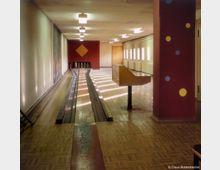 Ein schmaler Kegelbahnraum mit glänzendem Parkettboden und zwei Kegelbahnen, die zu einer rot gestrichenen Wand mit einem orangefarbenen Dekor führen. An der rechten Seite befinden sich Fensterreihen und Heizkörper, während links eine helle Wand verläuft; im Vordergrund steht ein hölzernes Bedienpult.