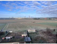 Weitläufige, frostbedeckte Ackerlandschaft mit einem kleinen, eingezäunten Hofgebäude im Vordergrund und industriellen Schornsteinen, aus denen Rauch aufsteigt, im Hintergrund. Der Himmel ist teils bewölkt mit blauen Abschnitten, und am Horizont sind Windräder und Strommasten zu erkennen.
