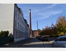 Straßenszene mit Kopfsteinpflaster und Straßenbahngleisen, flankiert von einem Mehrfamilienhaus links und einer Reihe von Bäumen rechts. Im Hintergrund befindet sich ein Backsteingebäude mit einem hohen Industrie-Schornstein.