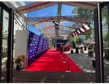 Ein überdachter Eingangsbereich mit einem roten Teppich, flankiert von Absperrungen und Beleuchtungseinrichtungen sowie einer dunkelblauen Wand mit Logos. Im Hintergrund ist ein Platz mit Bäumen und Gebäuden erkennbar.