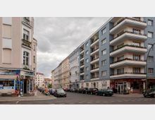 Straßenszene in einer Stadt mit mehrstöckigen Wohnhäusern; links ein beige Gebäude mit einem Internetcafé im Erdgeschoss, rechts ein moderneres blau-graues Wohnhaus mit Balkonen. Geparkte Autos stehen entlang der Straße, die bei bewölktem Himmel ruhig wirkt.